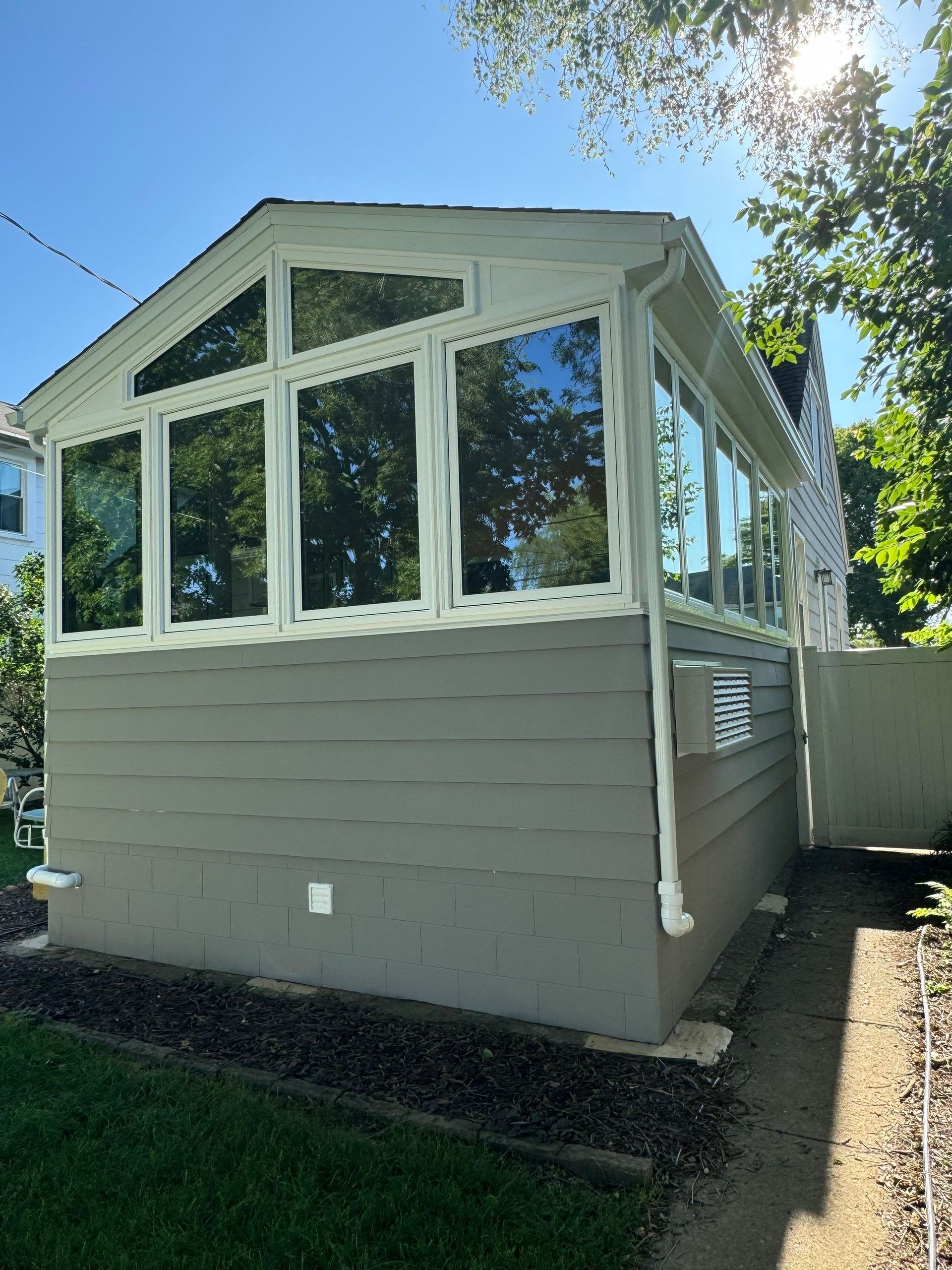 White-trimmed enclosed porch with many windows on a gray house, viewed from the backyard.