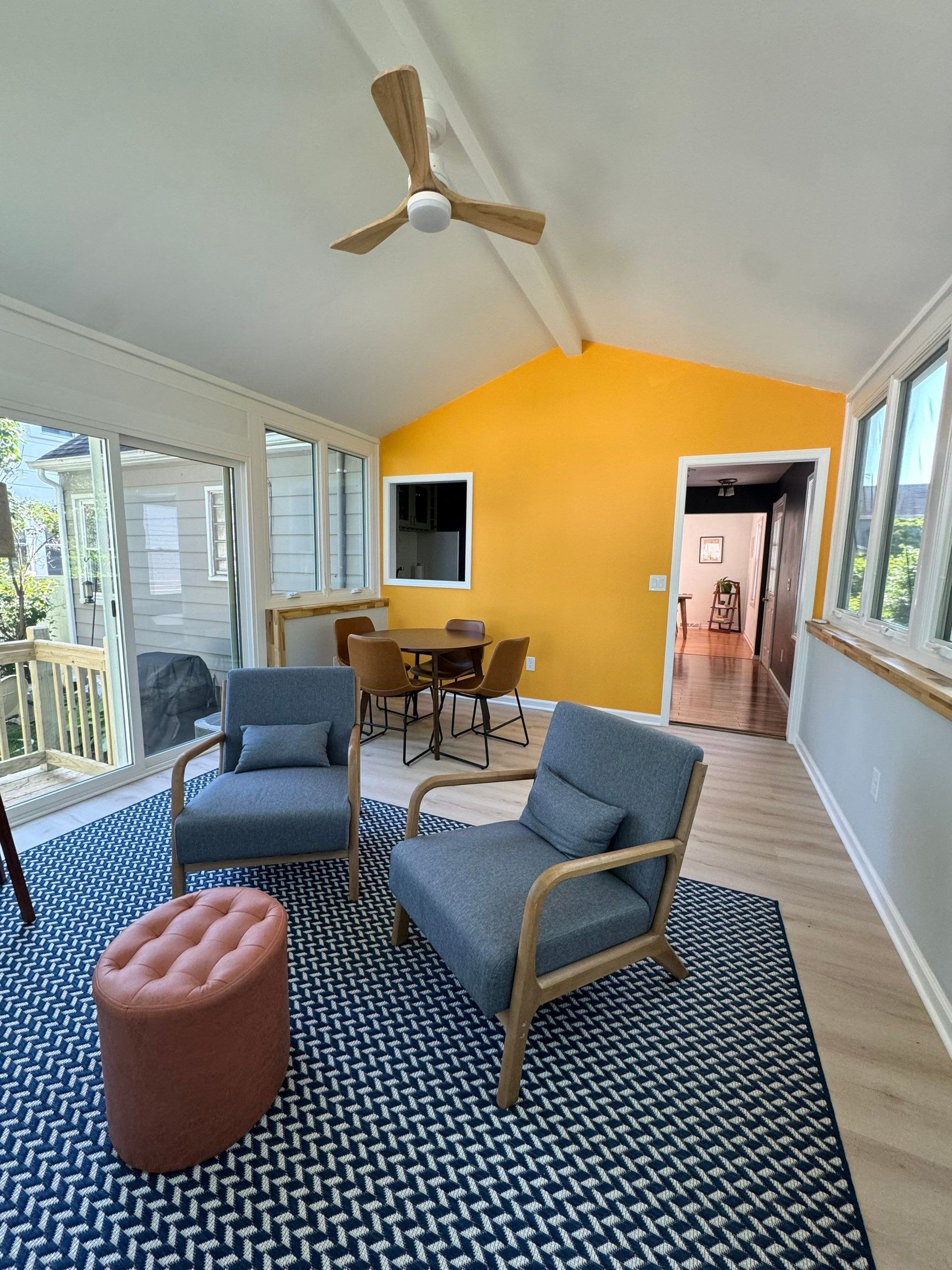 Sunroom with gray chairs, patterned rug, round ottoman, and yellow back wall under a ceiling fan.
