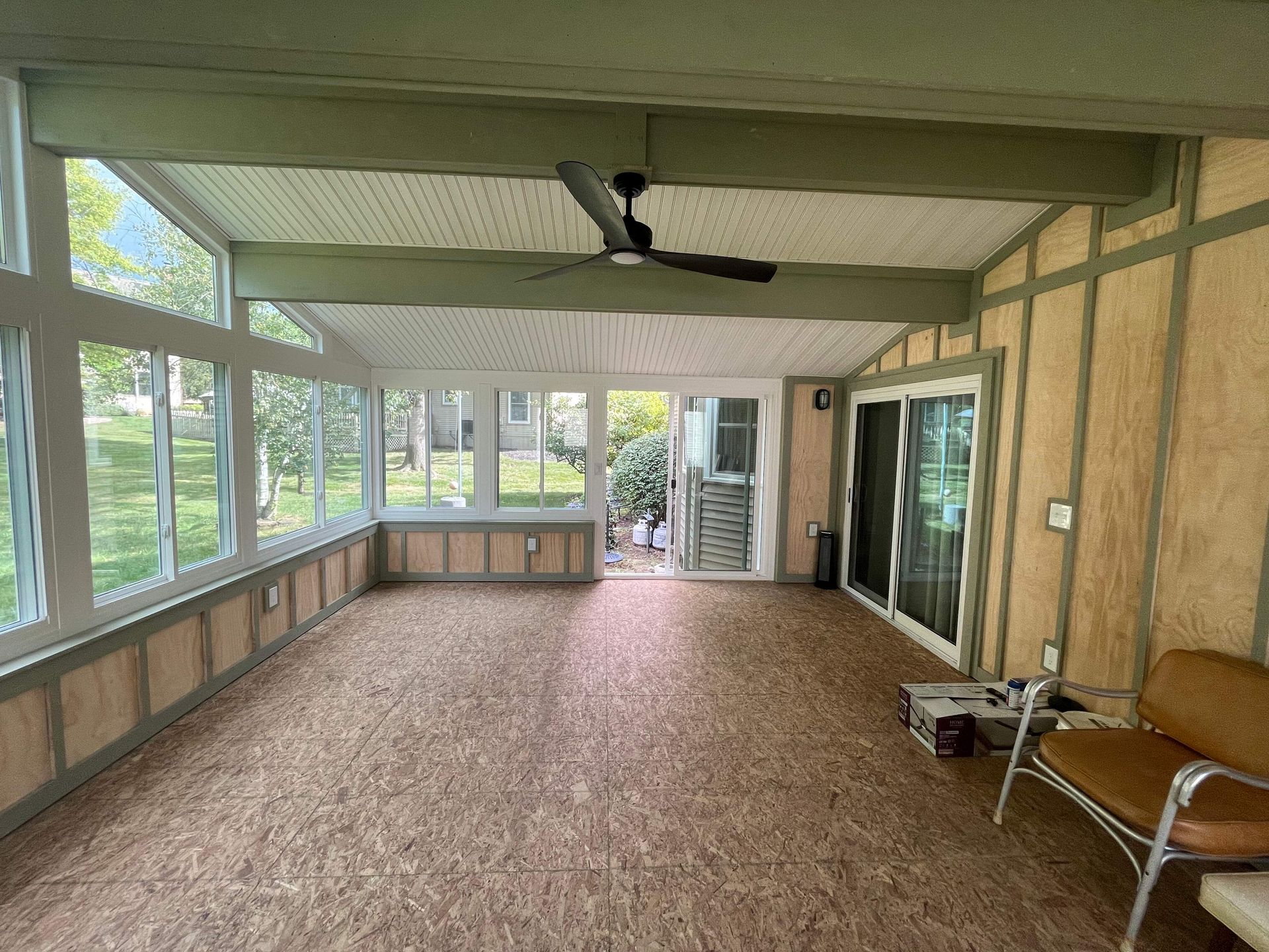 Empty screened-in porch with tan walls, brown carpet, large windows, and a chair near the right side.