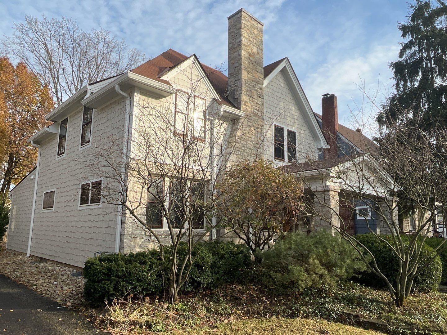 White house with stone chimney and dark roof, partly obscured by leafless trees and shrubs in a yard
