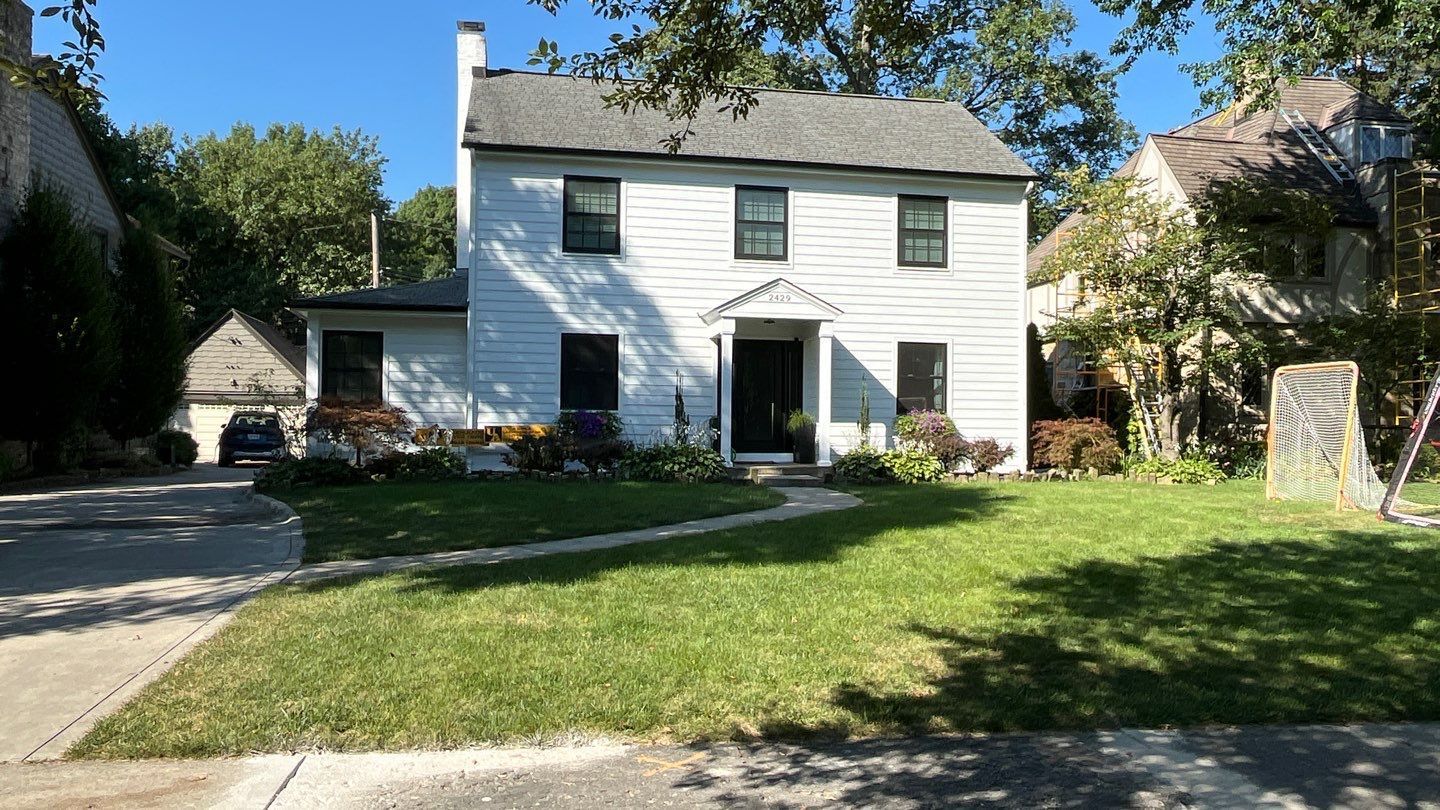White two-story house with a front lawn, driveway, and trees on a sunny day