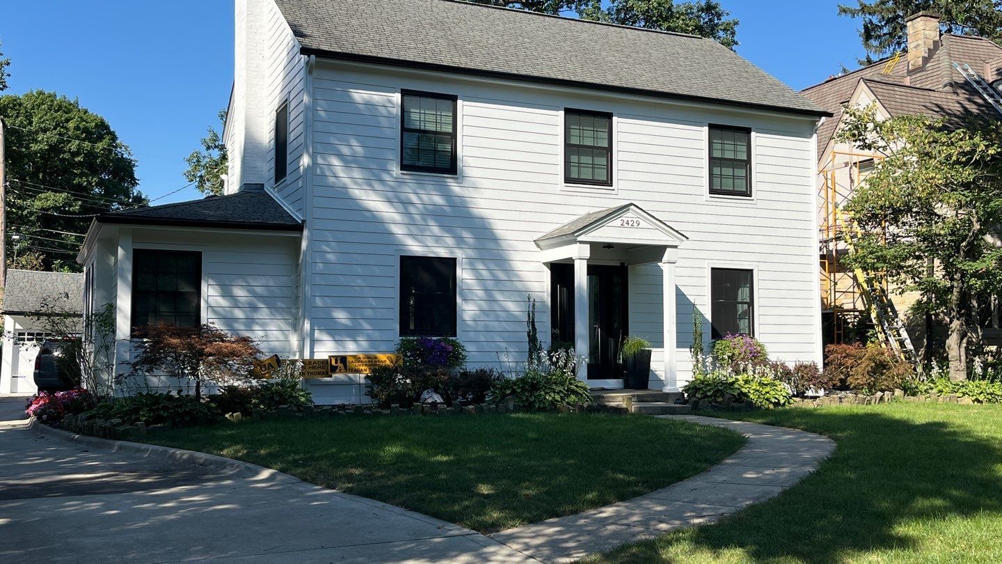 White two-story house with green shutters, front porch, and driveway on a sunny day