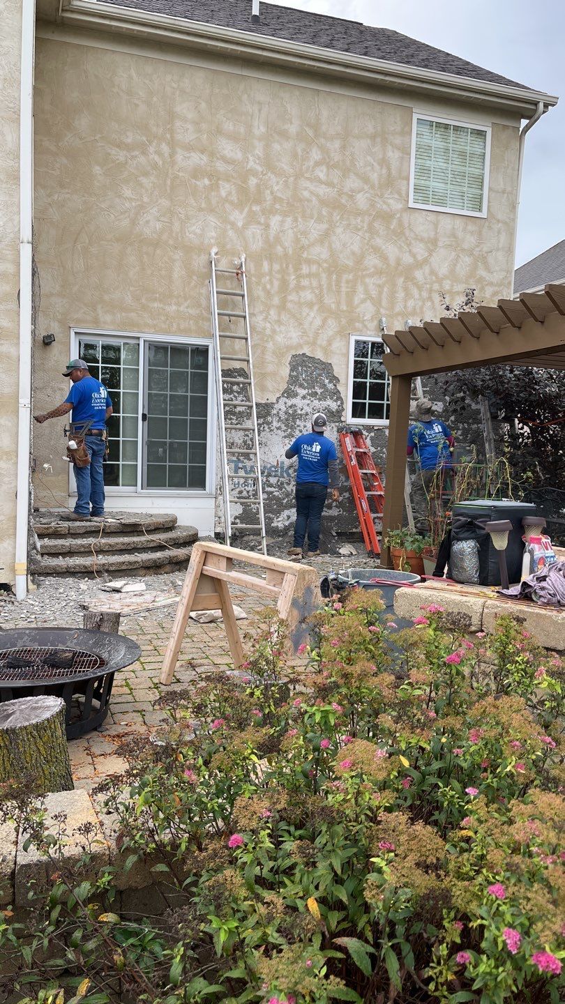 Two workers painting a beige house exterior, one on a ladder, with garden plants in the foreground.