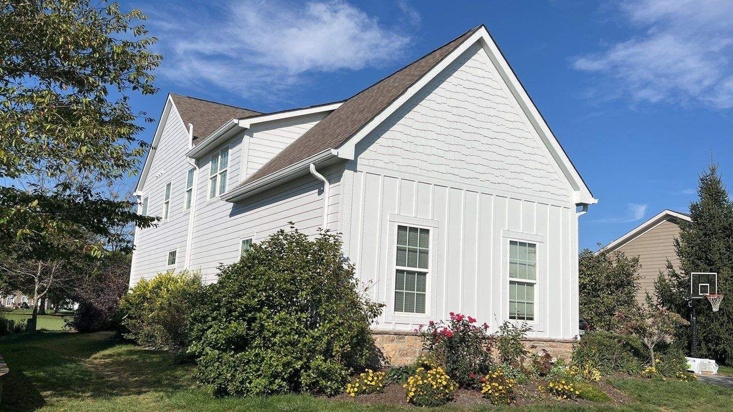 White house with a steep roof and green shutters, surrounded by trees and shrubs under a blue sky