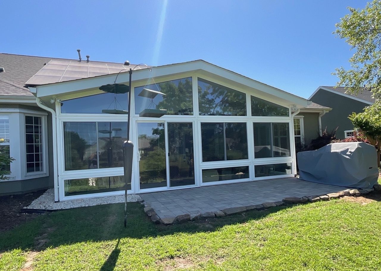 Sunroom with large glass windows and white trim attached to a house, next to a grassy yard.