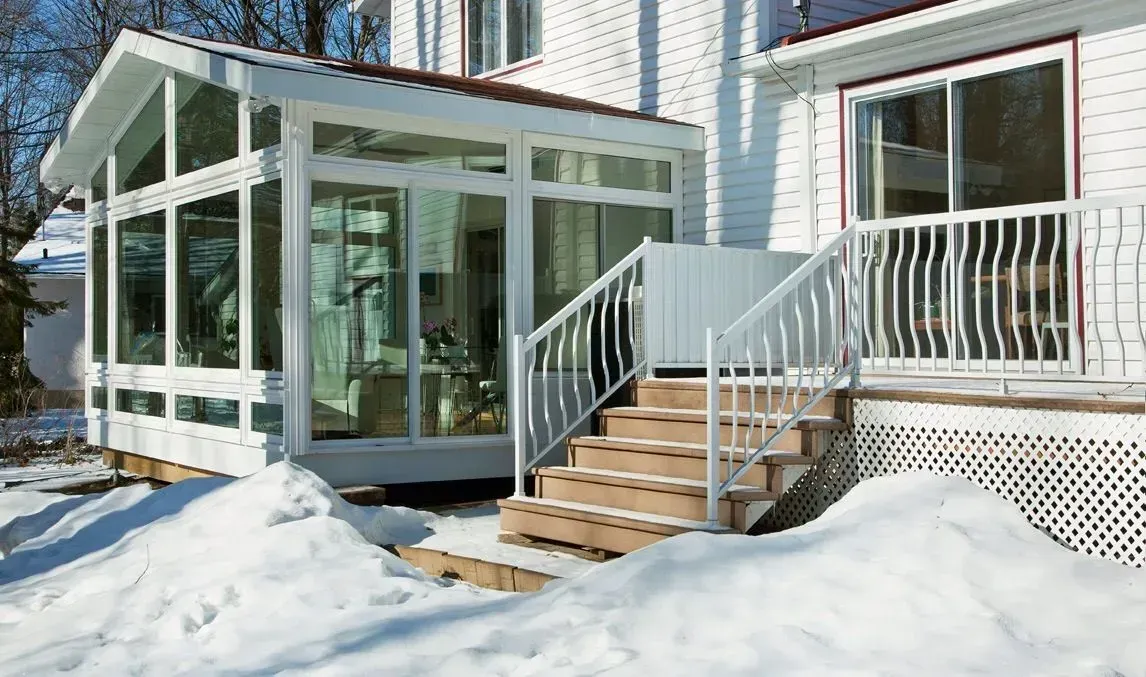Snowy house entrance with a glass sunroom, stairs, and white porch railing