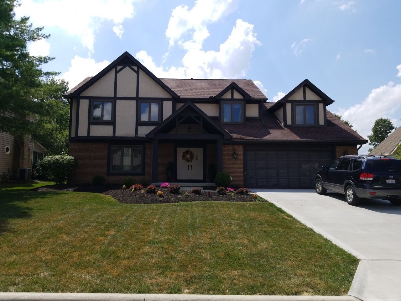 Suburban Tudor-style house with a front lawn, driveway, and parked SUV on a sunny day