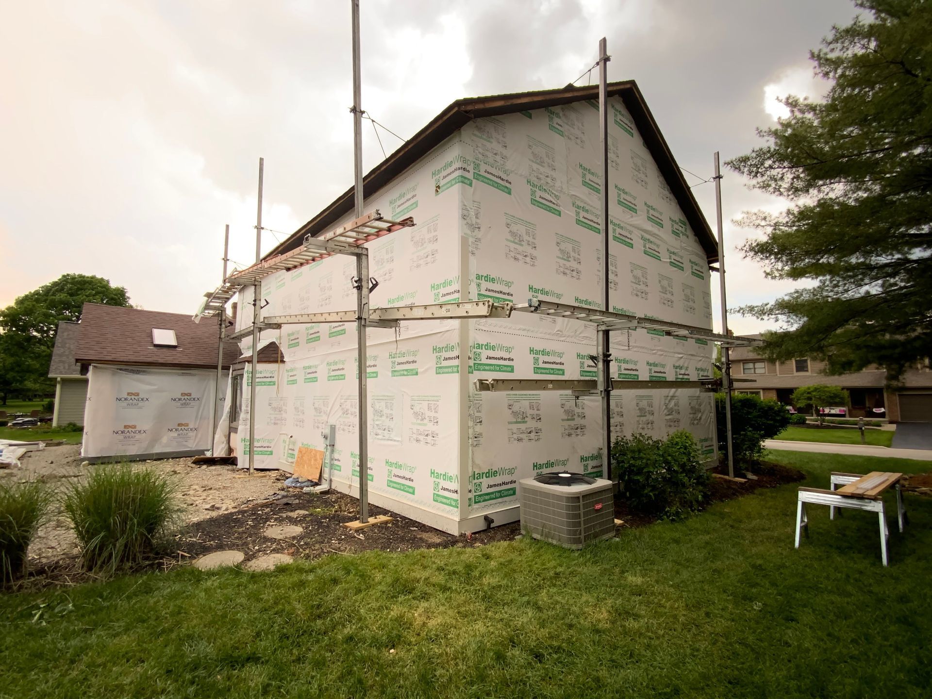 House under construction wrapped in white house wrap, with scaffolding poles and a yard in front