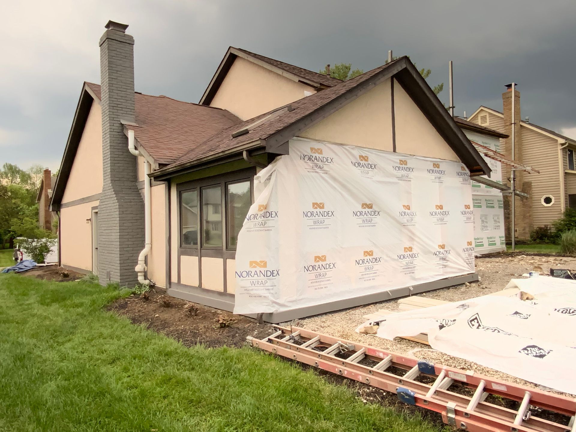 House under renovation with white house wrap, exposed roof framing, and ladder on a grassy lot