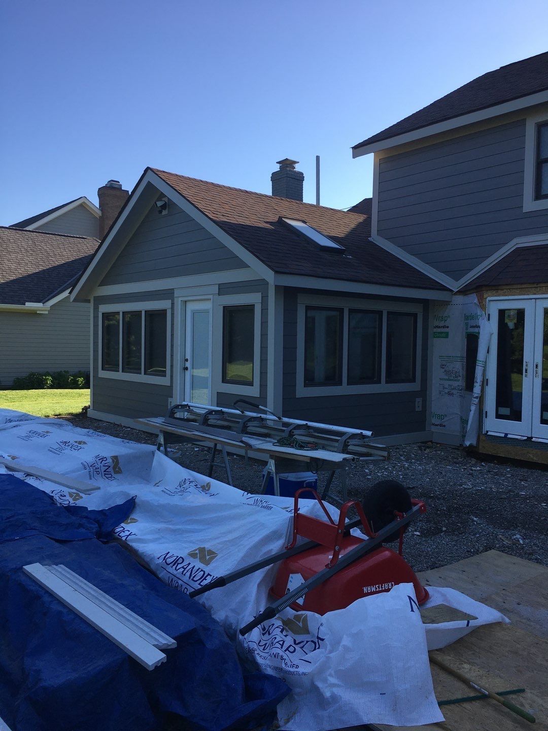 House exterior with white tarp, debris, and a red wheelbarrow in the yard under a blue sky