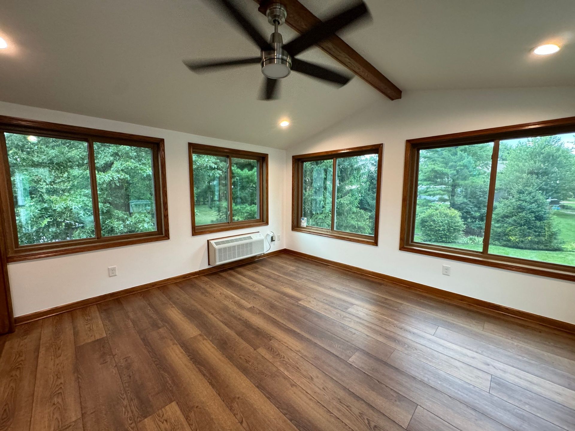 Empty sunlit room with wood floors, large windows, and a ceiling fan.