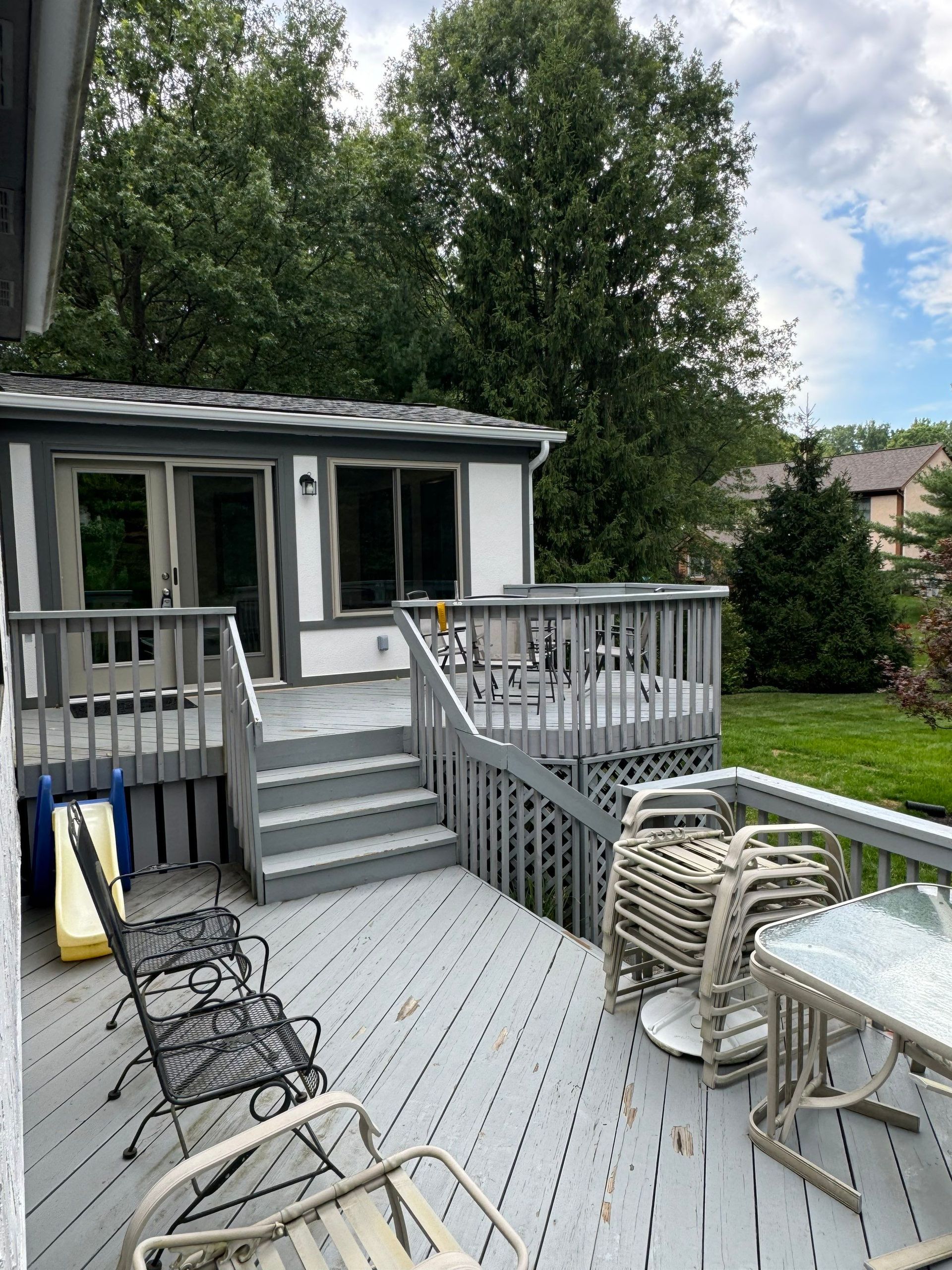 Back deck with gray railing, stairs, and outdoor chairs beside a white house and trees