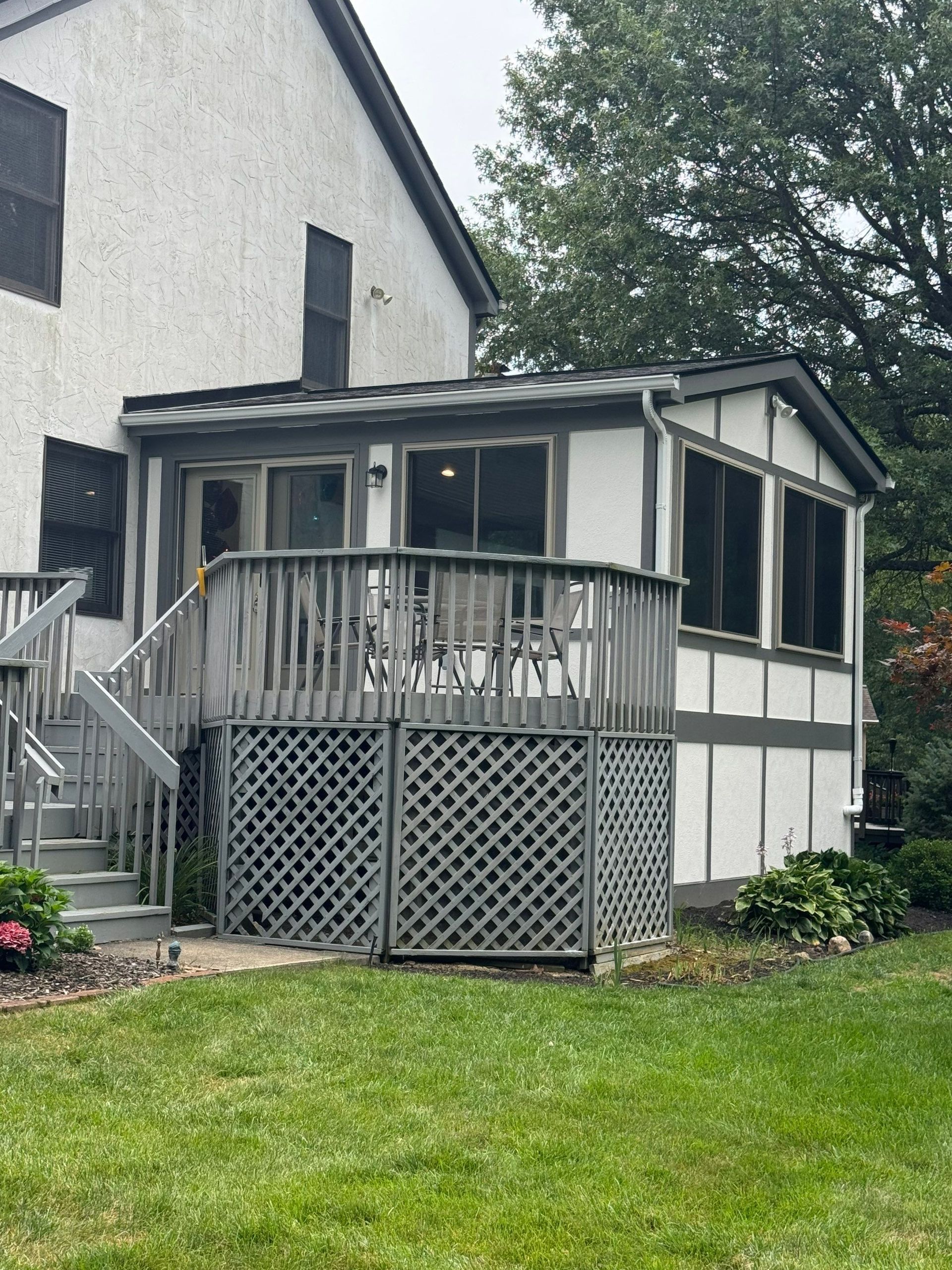 Small screened porch with gray railing and lattice on a house, beside a green lawn and trees