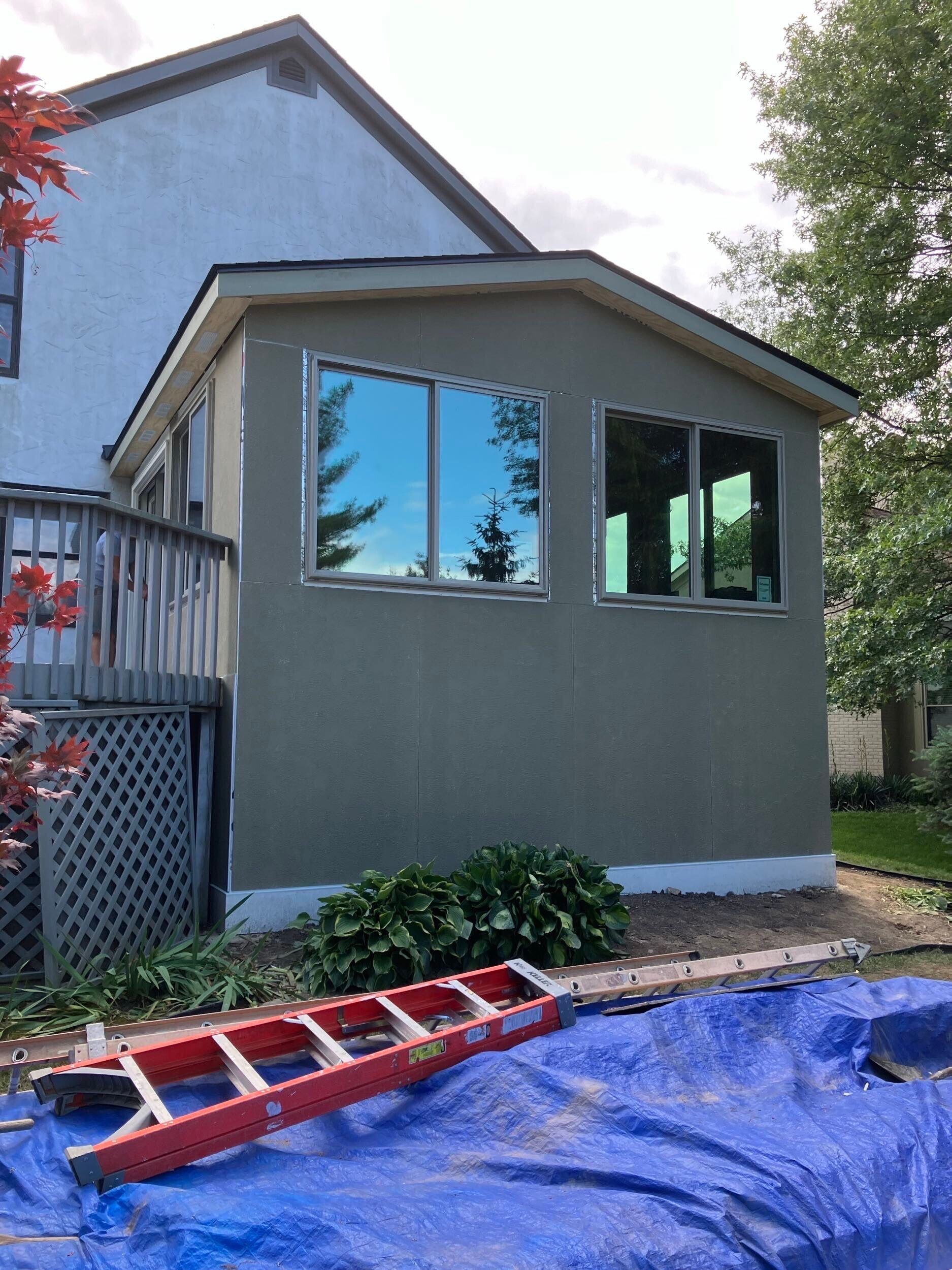 Small gray house addition with large windows, a red ladder, and a blue tarp in a yard