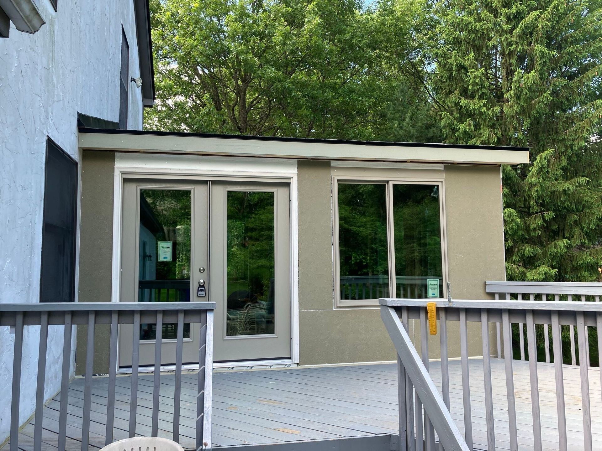 Small beige exterior room with glass doors, windows, and a gray deck railing surrounded by trees.