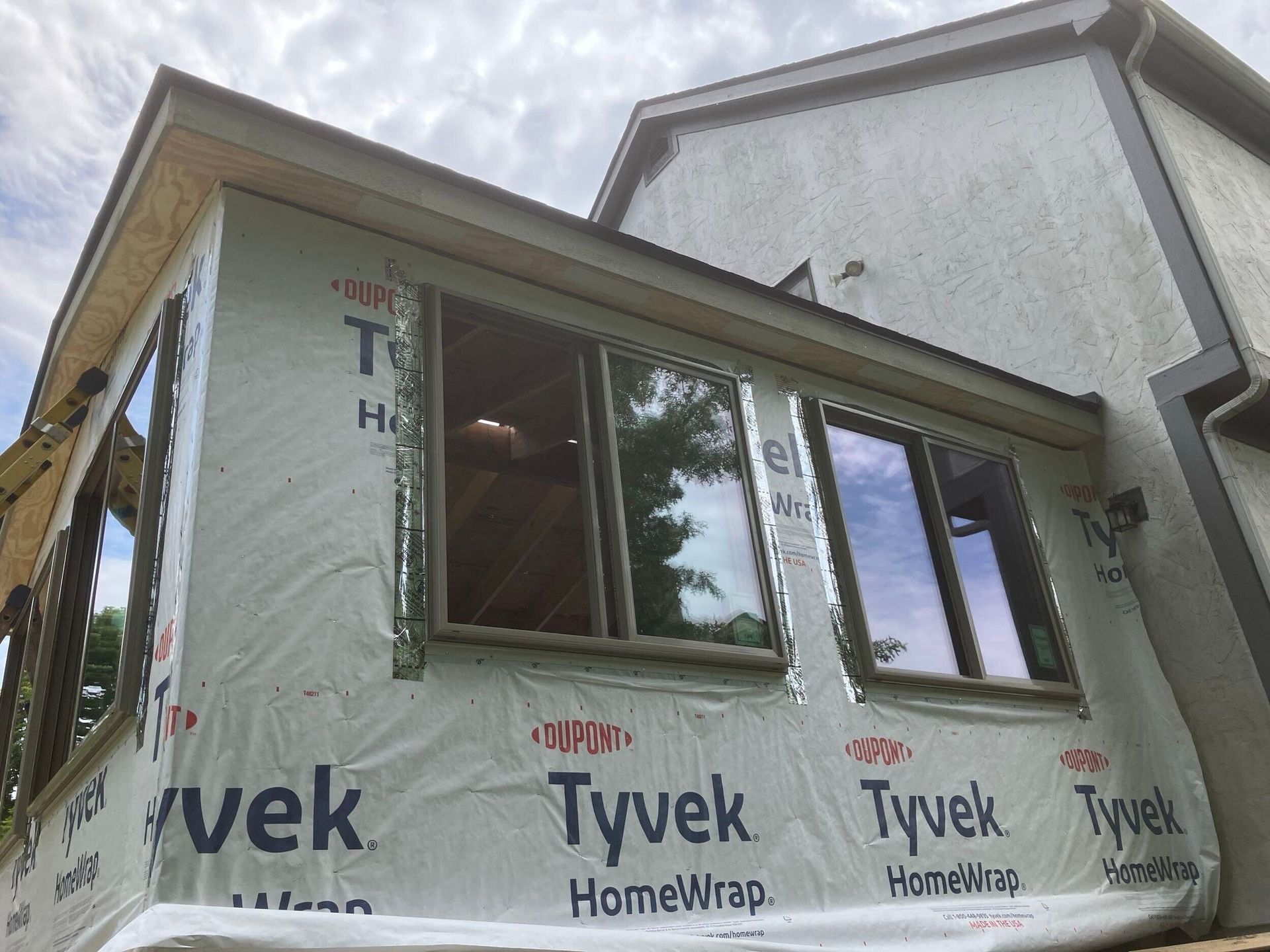 Partly wrapped house addition with Tyvek and two windows under an overcast sky