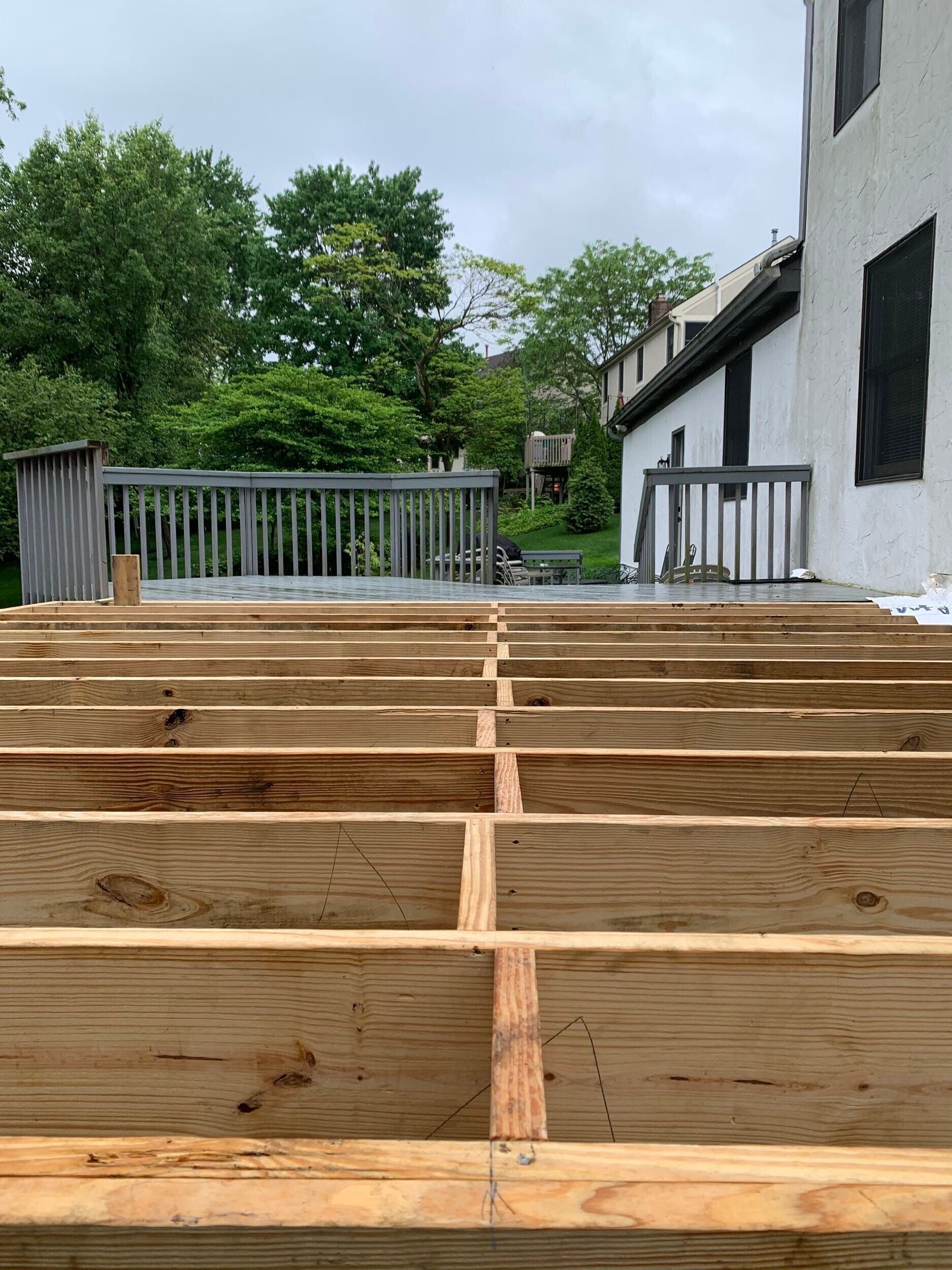 Wooden deck framing under construction beside a white building, with trees and a cloudy sky in the background