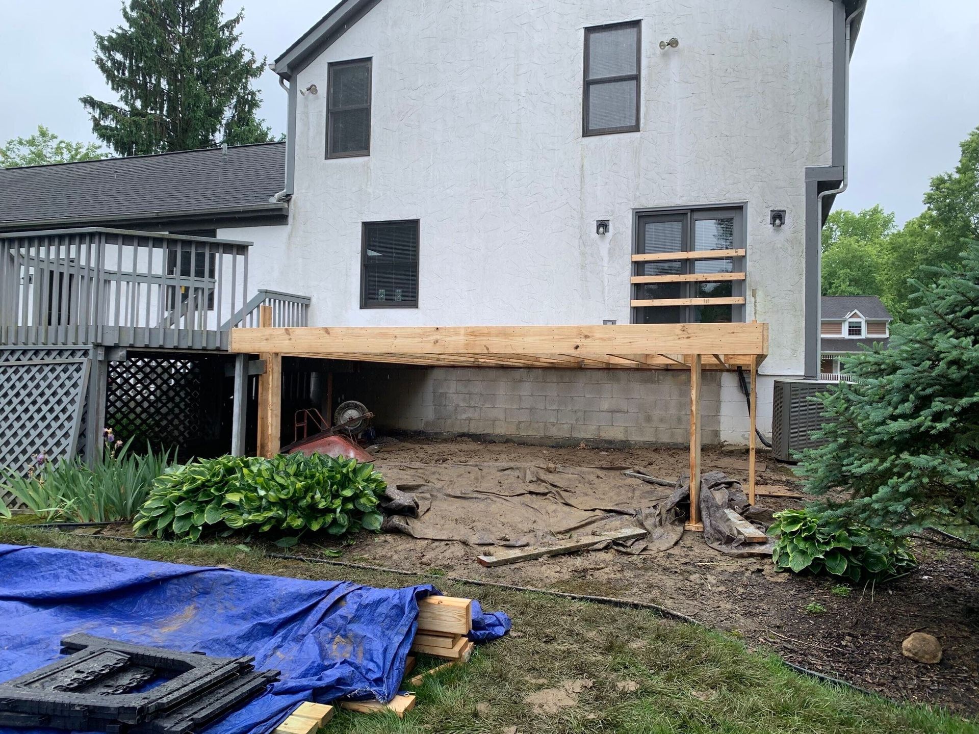 Backyard deck under construction beside a white house, with exposed framing and dirt ground.