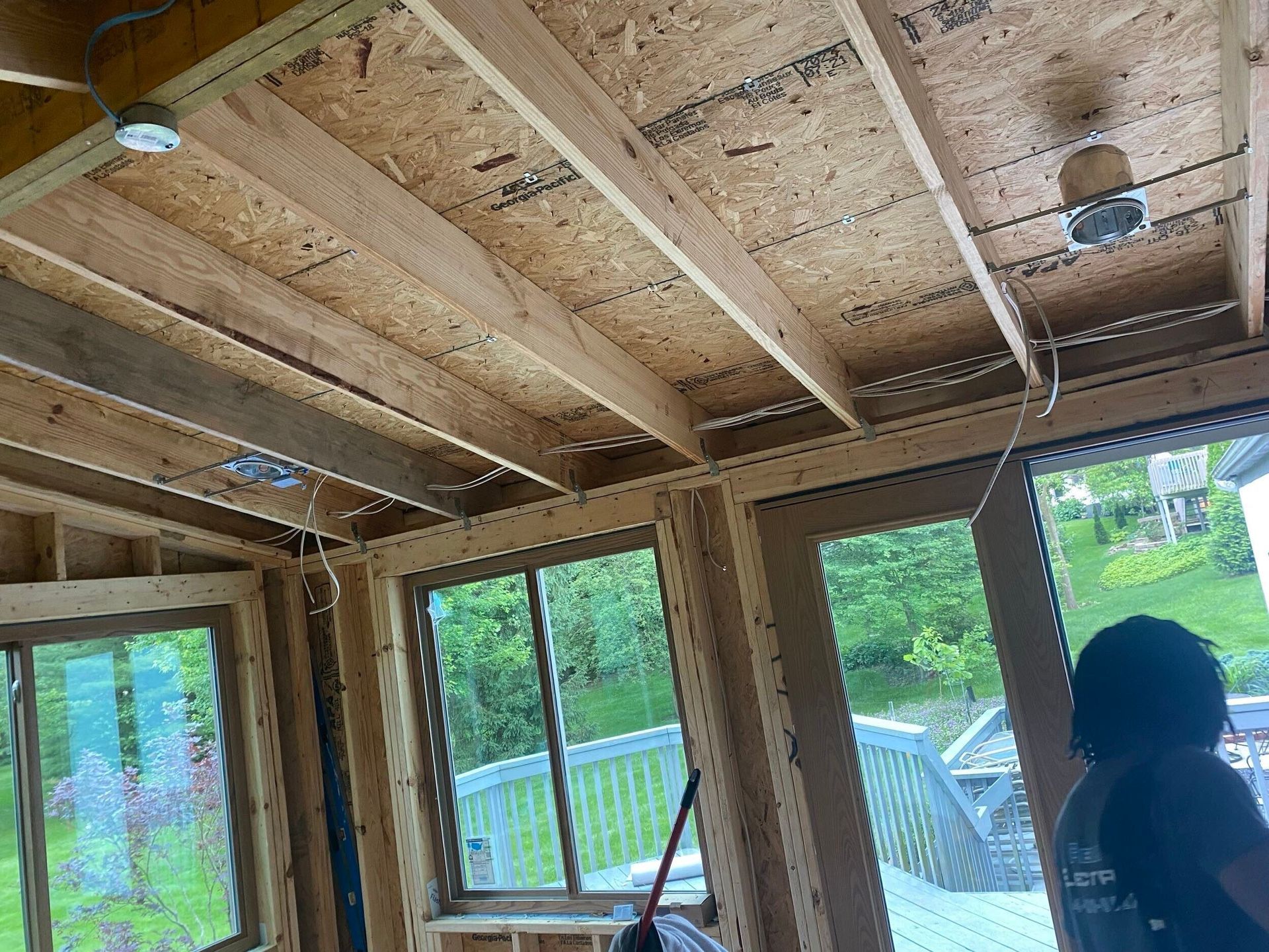 Exposed wooden ceiling framing inside a house under construction, with large windows and a person near the right edge.