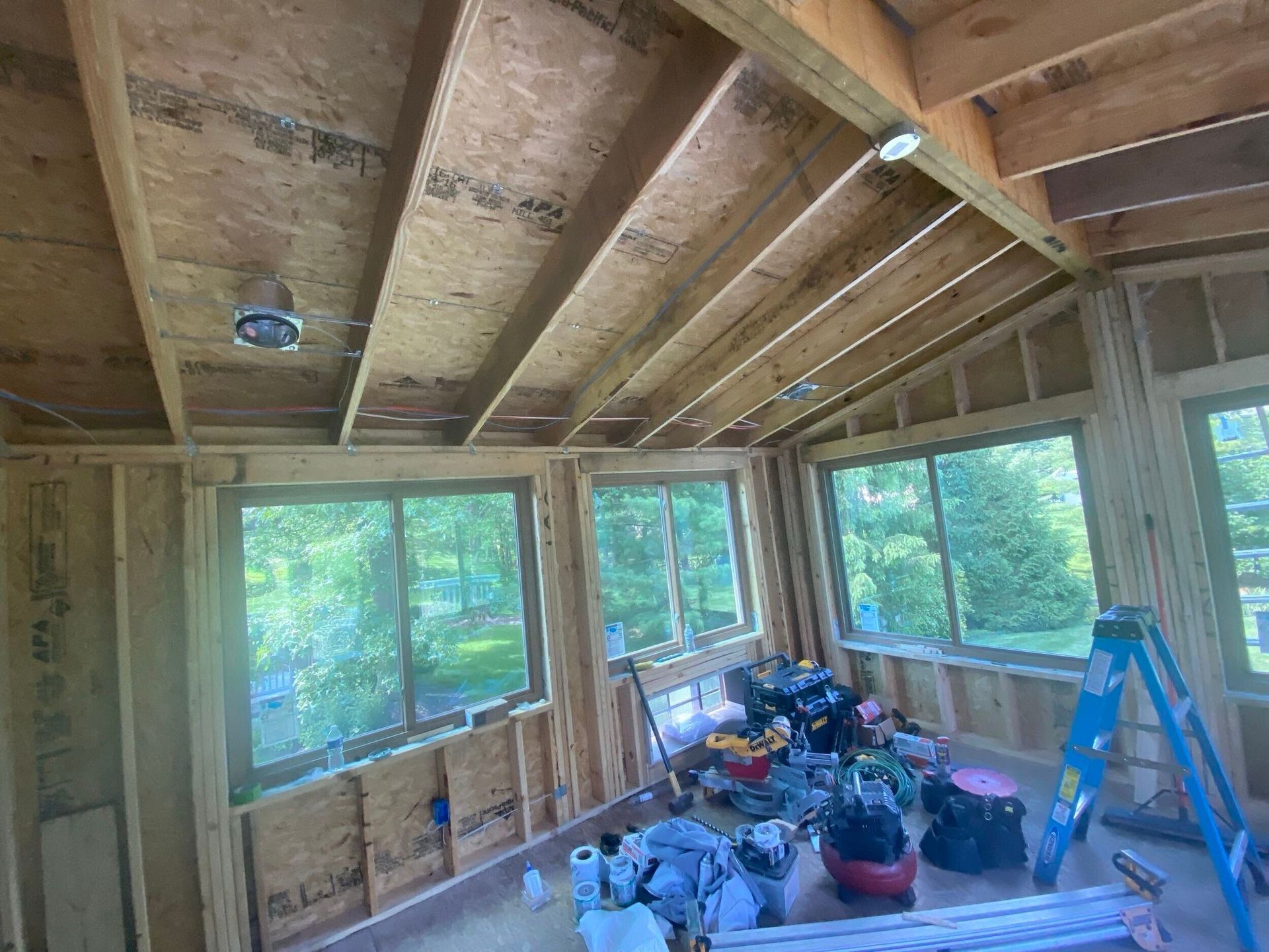Unfinished sunroom interior with exposed wood framing, windows, a ladder, and potted plants on the floor.