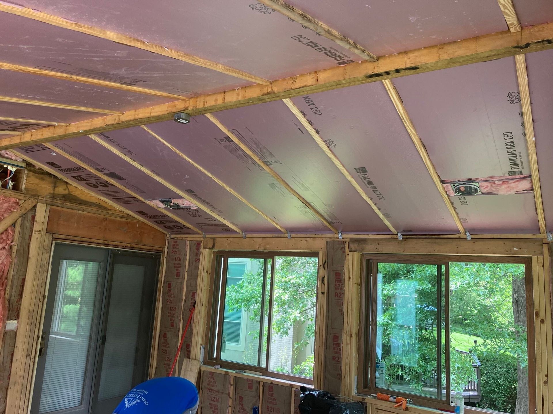 Unfinished room with exposed framing, purple ceiling insulation, and large windows looking onto green trees