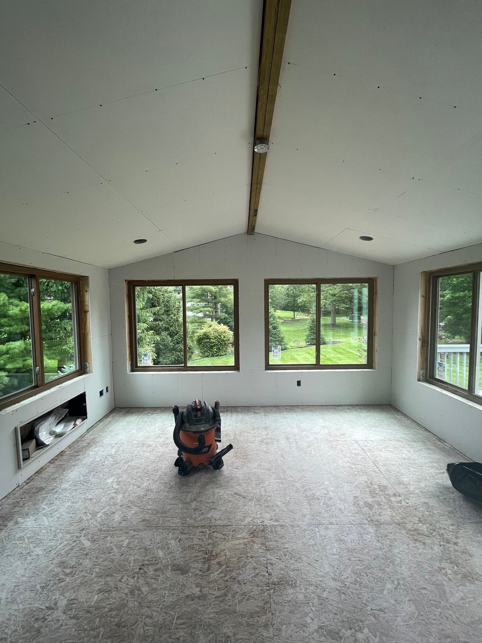 Empty bright room with large windows, white walls, exposed beam, and a vacuum on the floor
