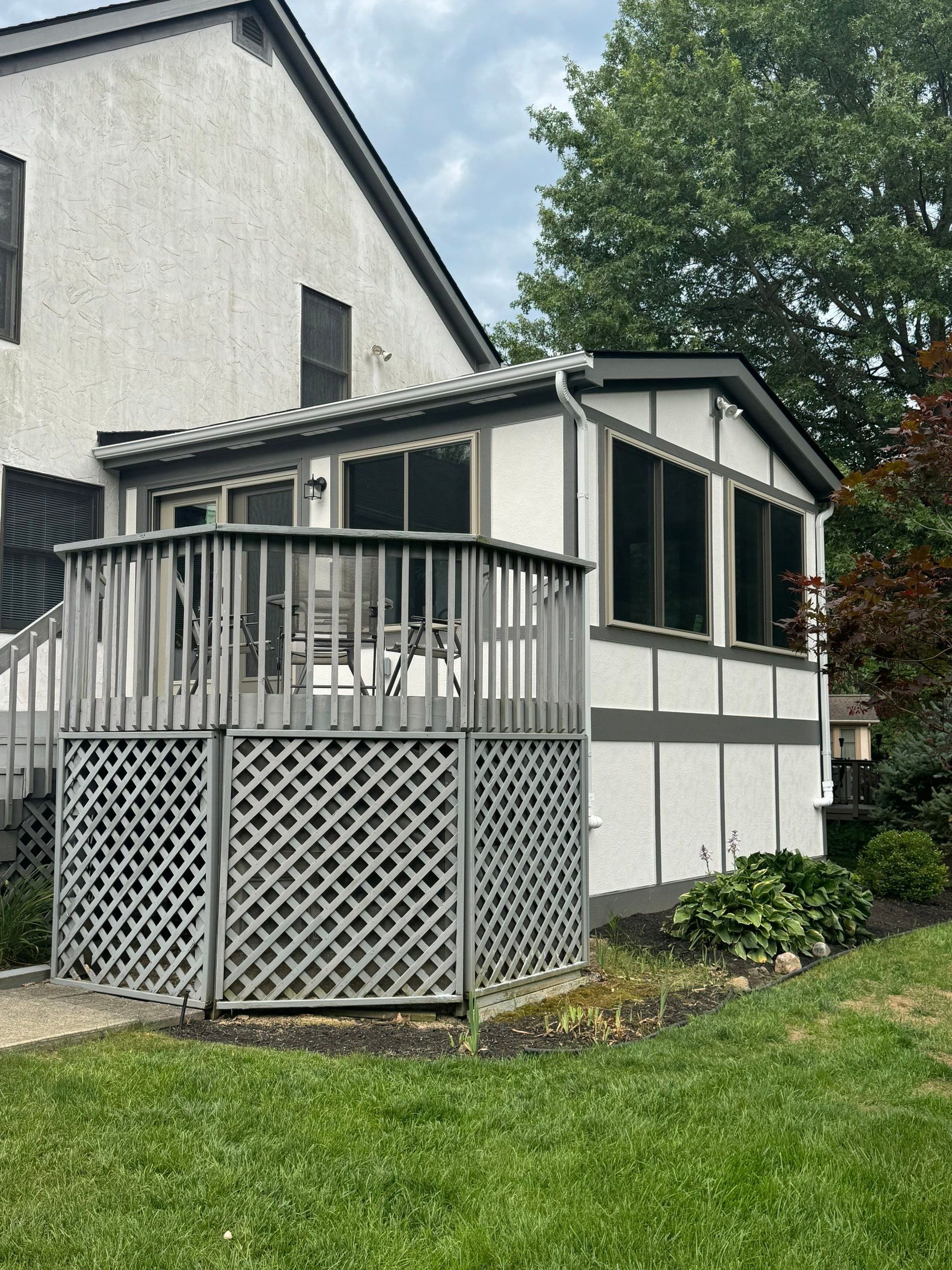 Enclosed white porch with lattice skirting and railing beside a gray house, overlooking a green lawn.