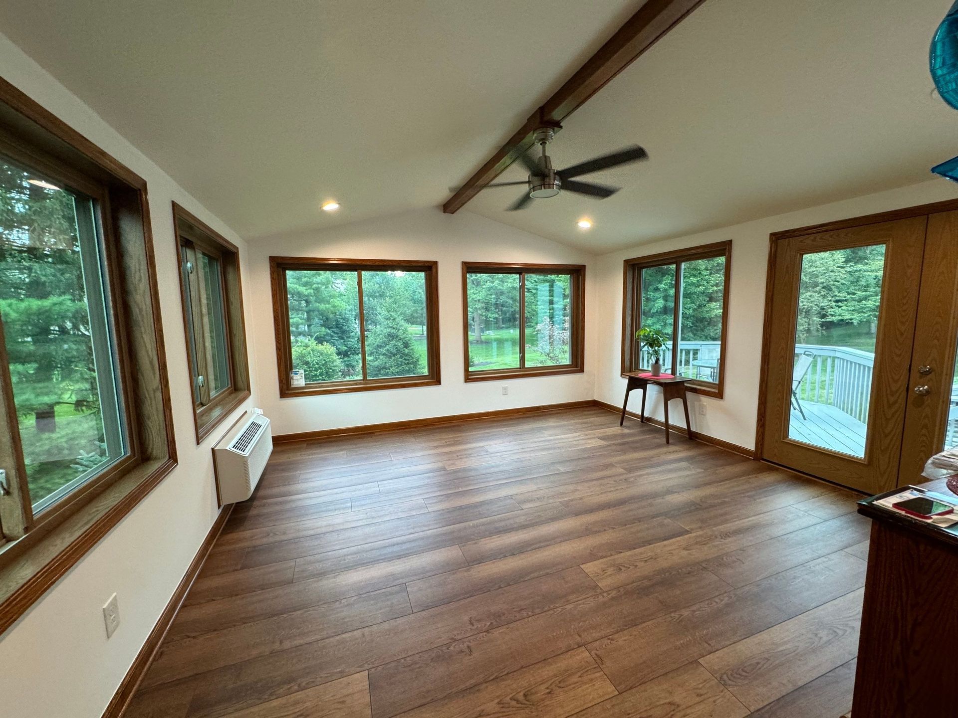 Empty sunlit living room with wood floor, large windows, and ceiling fan overlooking greenery