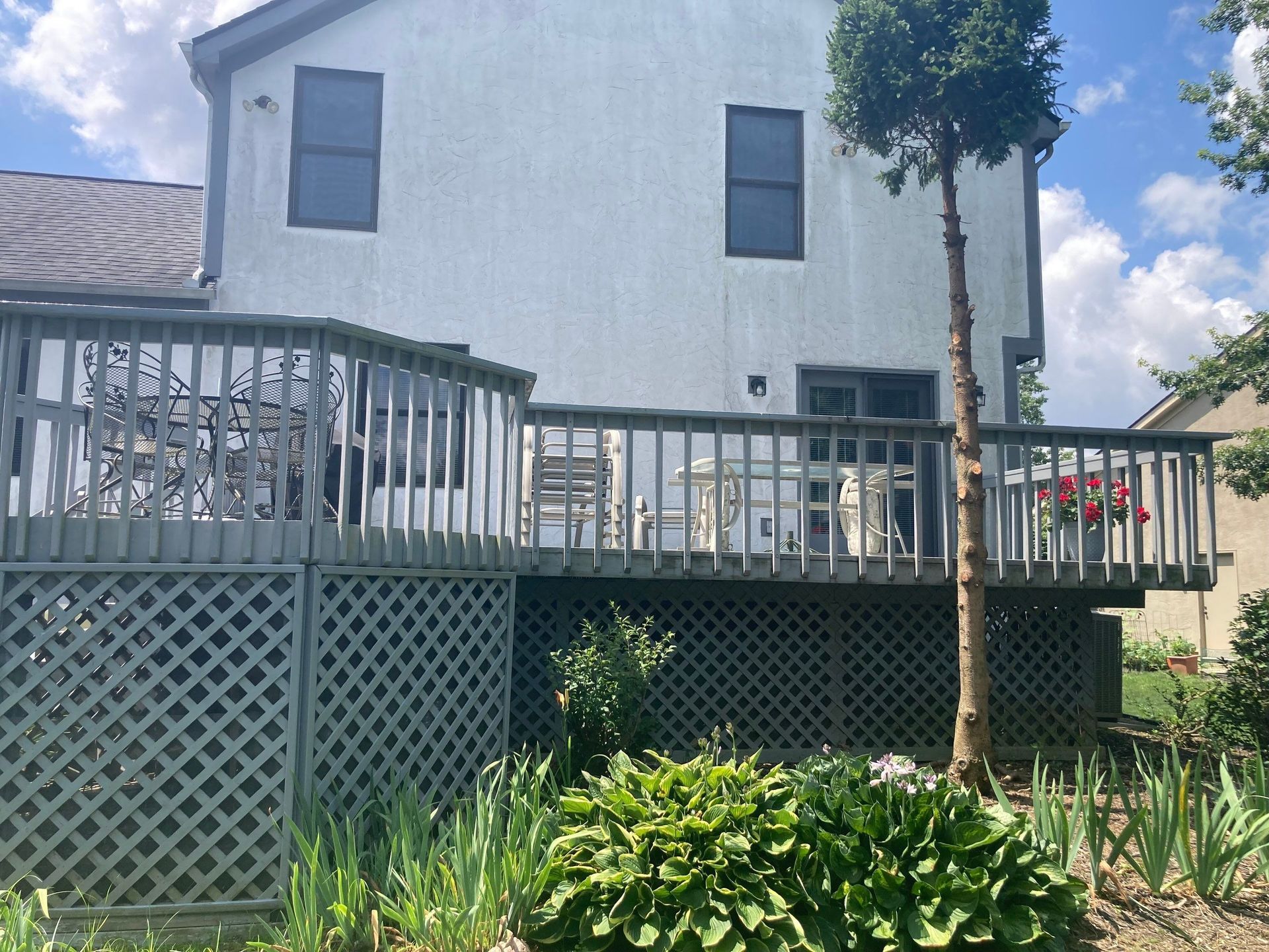 White house with gray deck railing, lattice skirting, and a small tree under a blue sky.