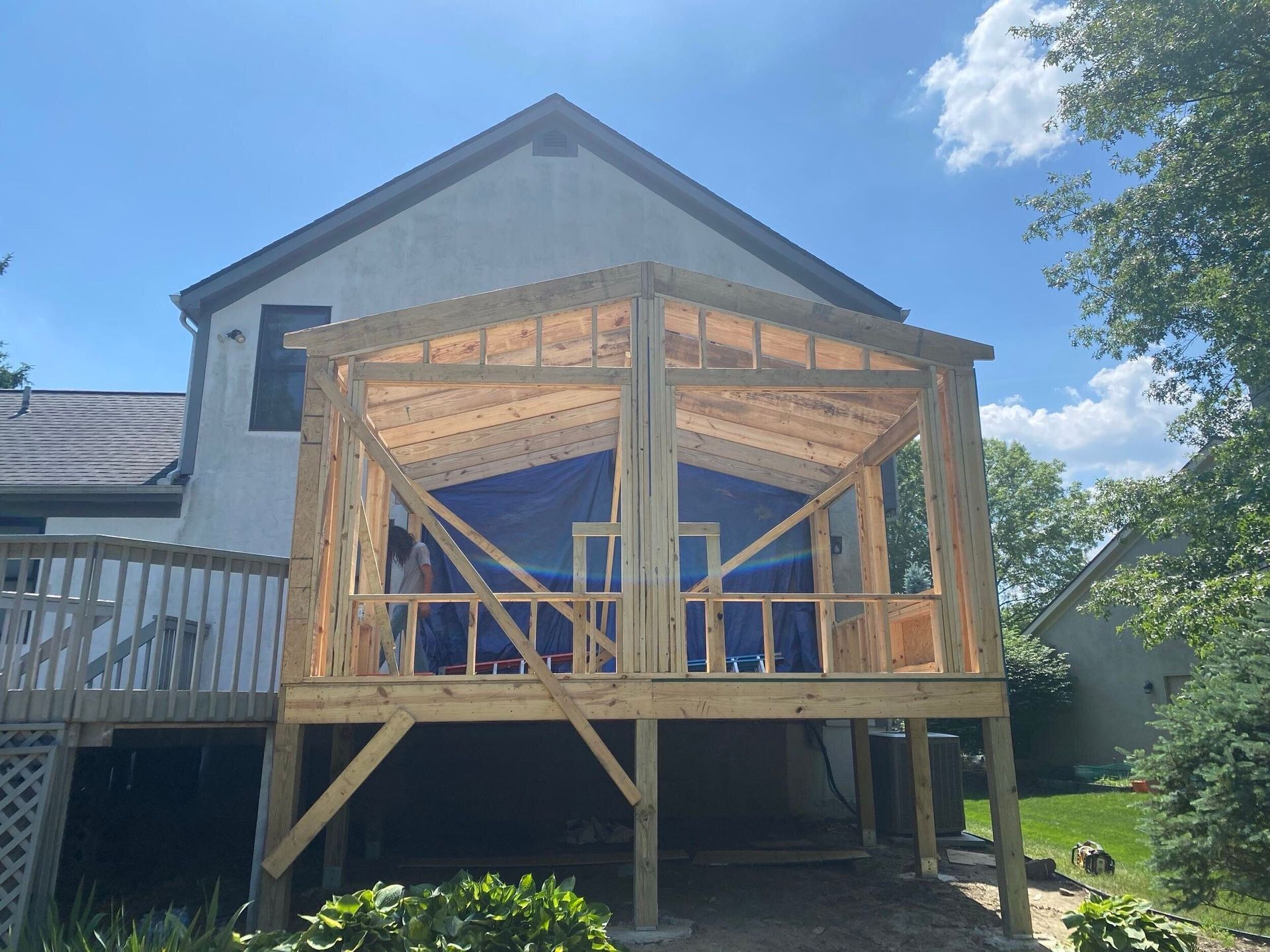 Partially built wooden porch addition on a house with exposed framing and a blue tarp behind it