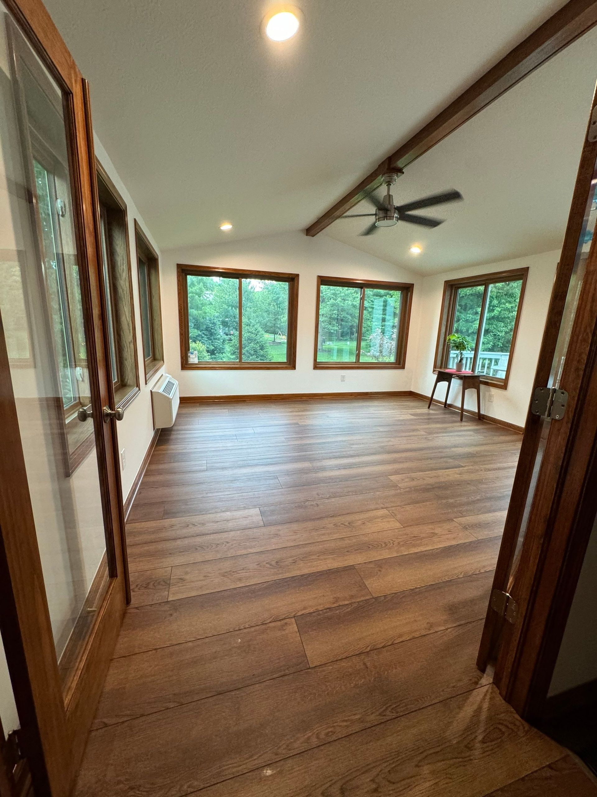 Empty sunroom with wood floors, recessed lights, ceiling fan, and large windows overlooking trees