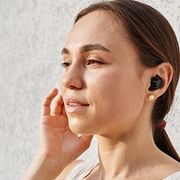 Woman wearing a black earbud, touching her face against a light background.