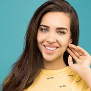 Smiling woman with long brown hair, touching her face against a teal background