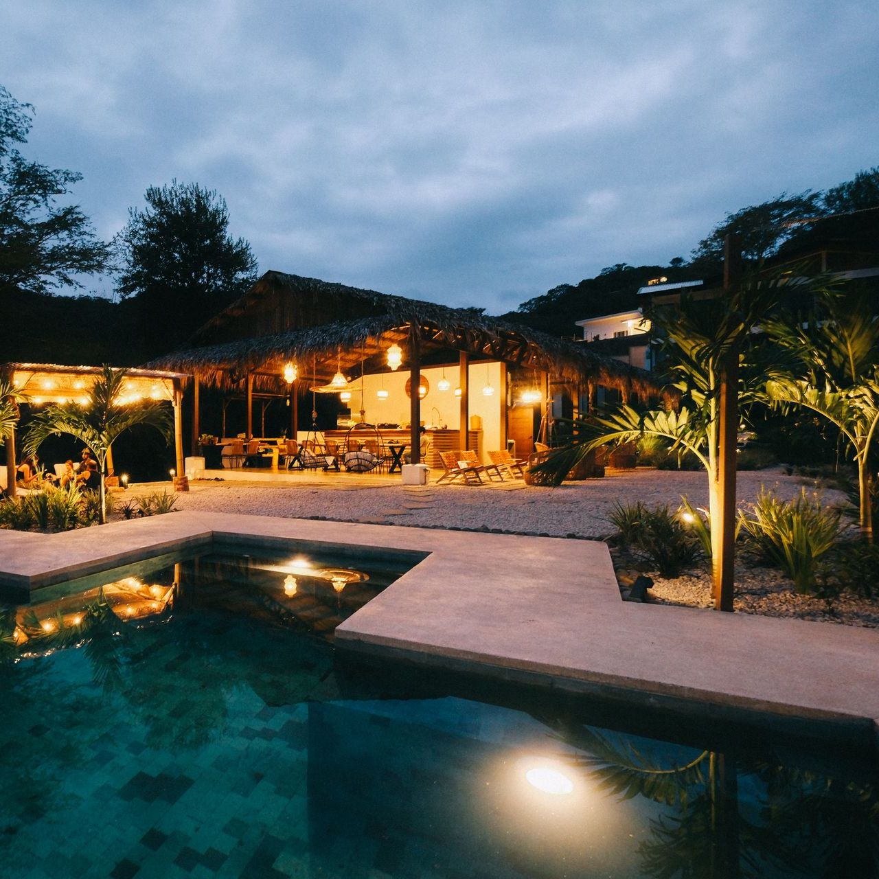 A tropical resort at dusk with a pool in the foreground and a thatched-roof building lit up in the background.