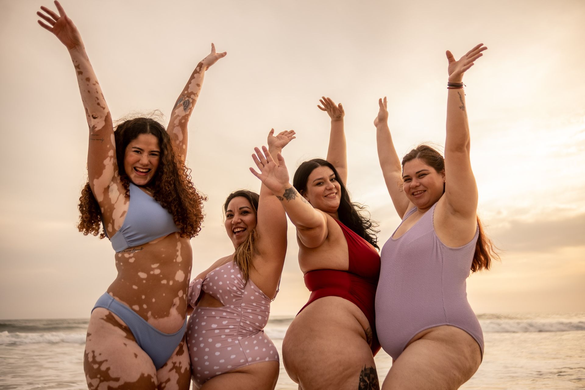 Four diverse women in swimwear on a beach, arms raised in celebration. Golden hour light, ocean in background.