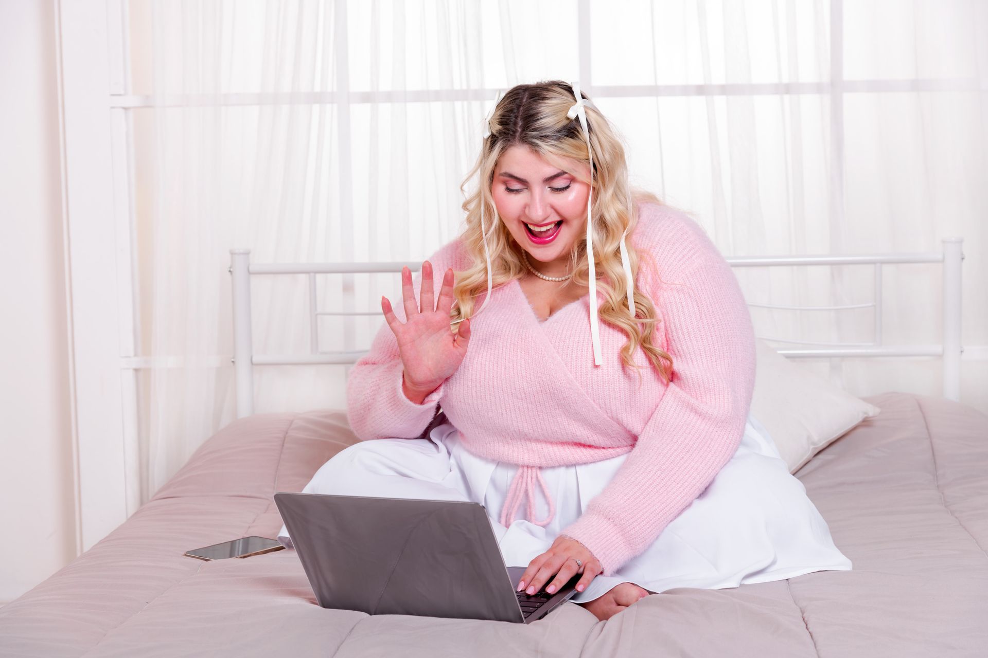 Blonde woman in pink sweater waves at a laptop screen while sitting on a bed in a bright room.