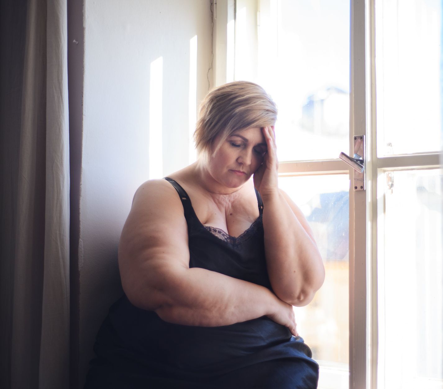 A woman with her hand on her forehead, appearing distressed, sits by a window. She is wearing a black dress.