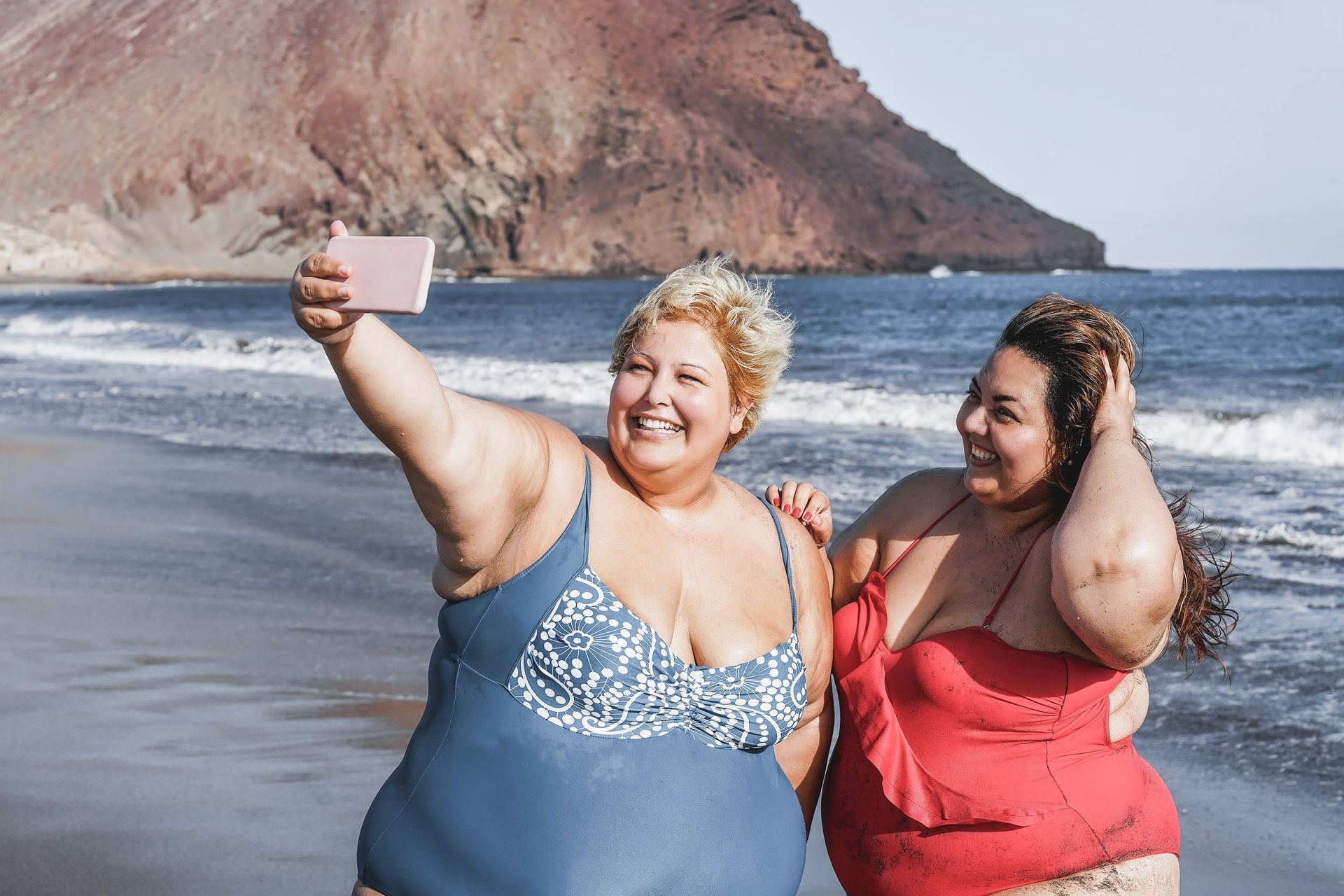 Two plus-size women on a beach, one taking a selfie with a phone. The woman in blue smiles, the other in red smiles too.