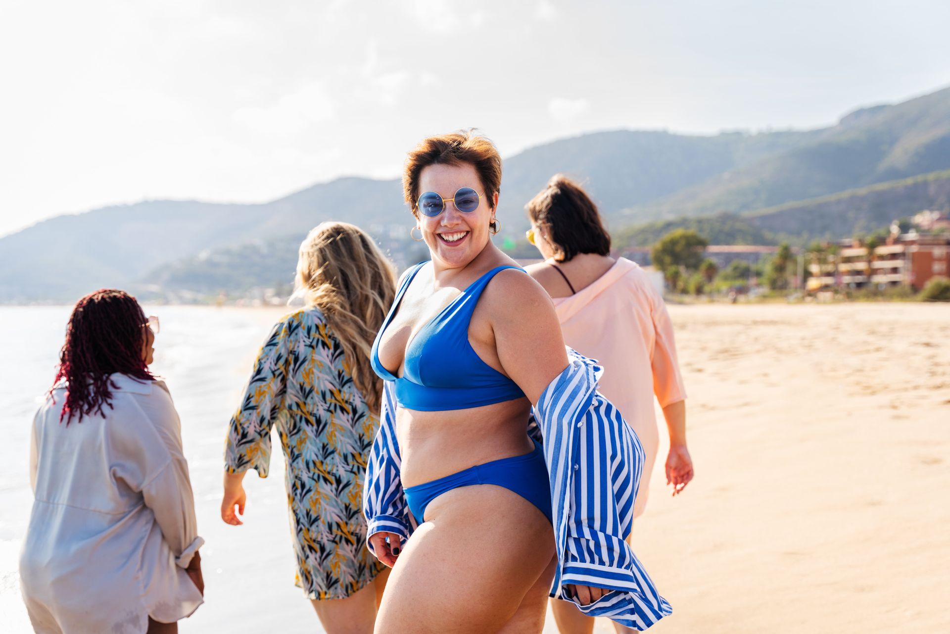 Four women on a beach: smiles, another in a patterned cover-up, and two others walk in the background.