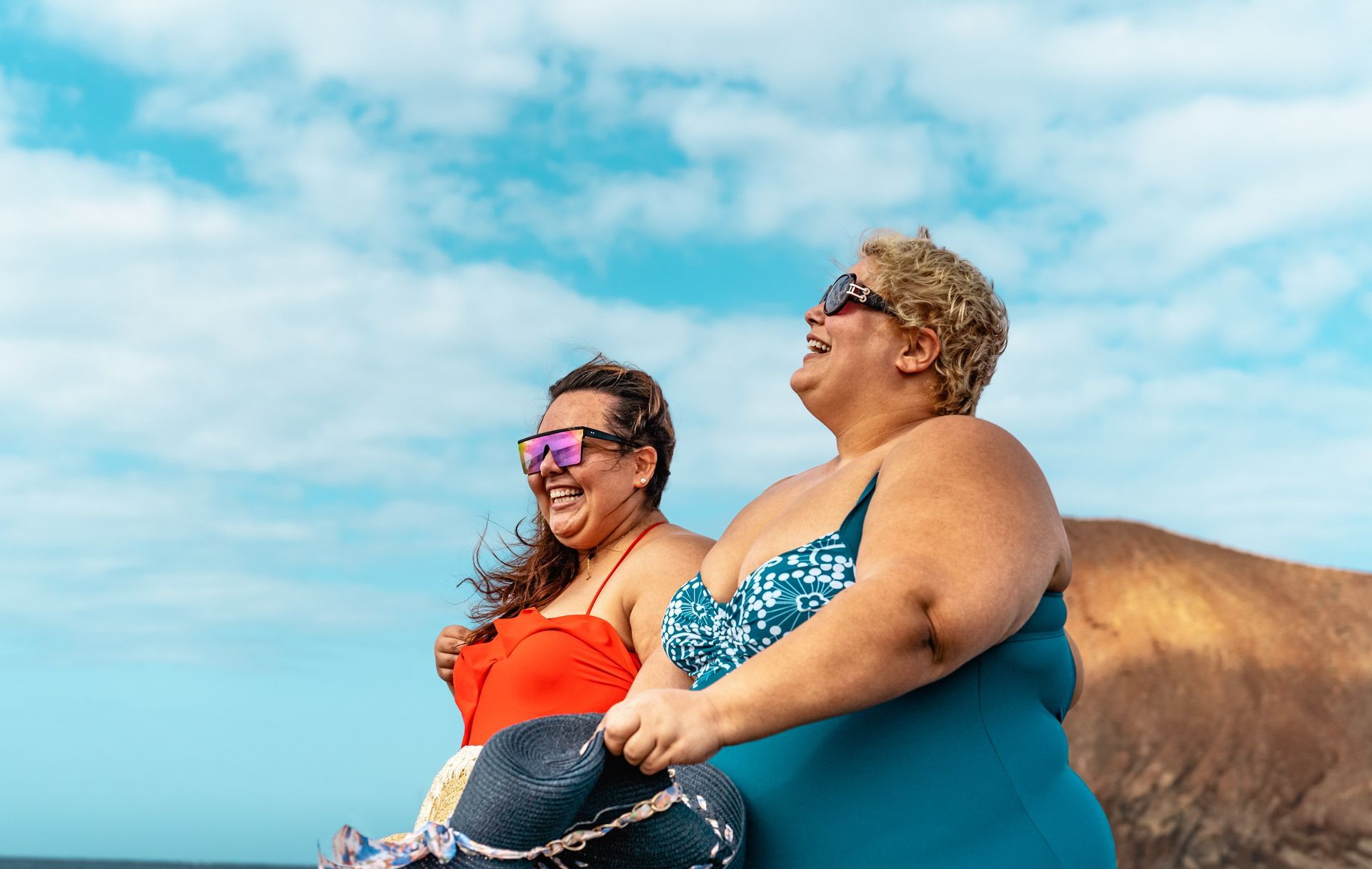 Two smiling women in swimwear, sunglasses, and a blue sky background.