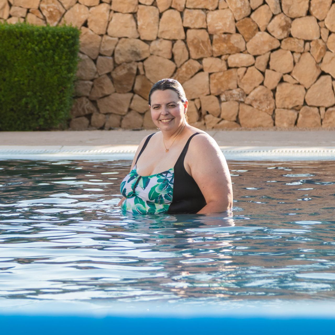 Woman in a black and green swimsuit standing in a pool, smiling. Stone wall background.