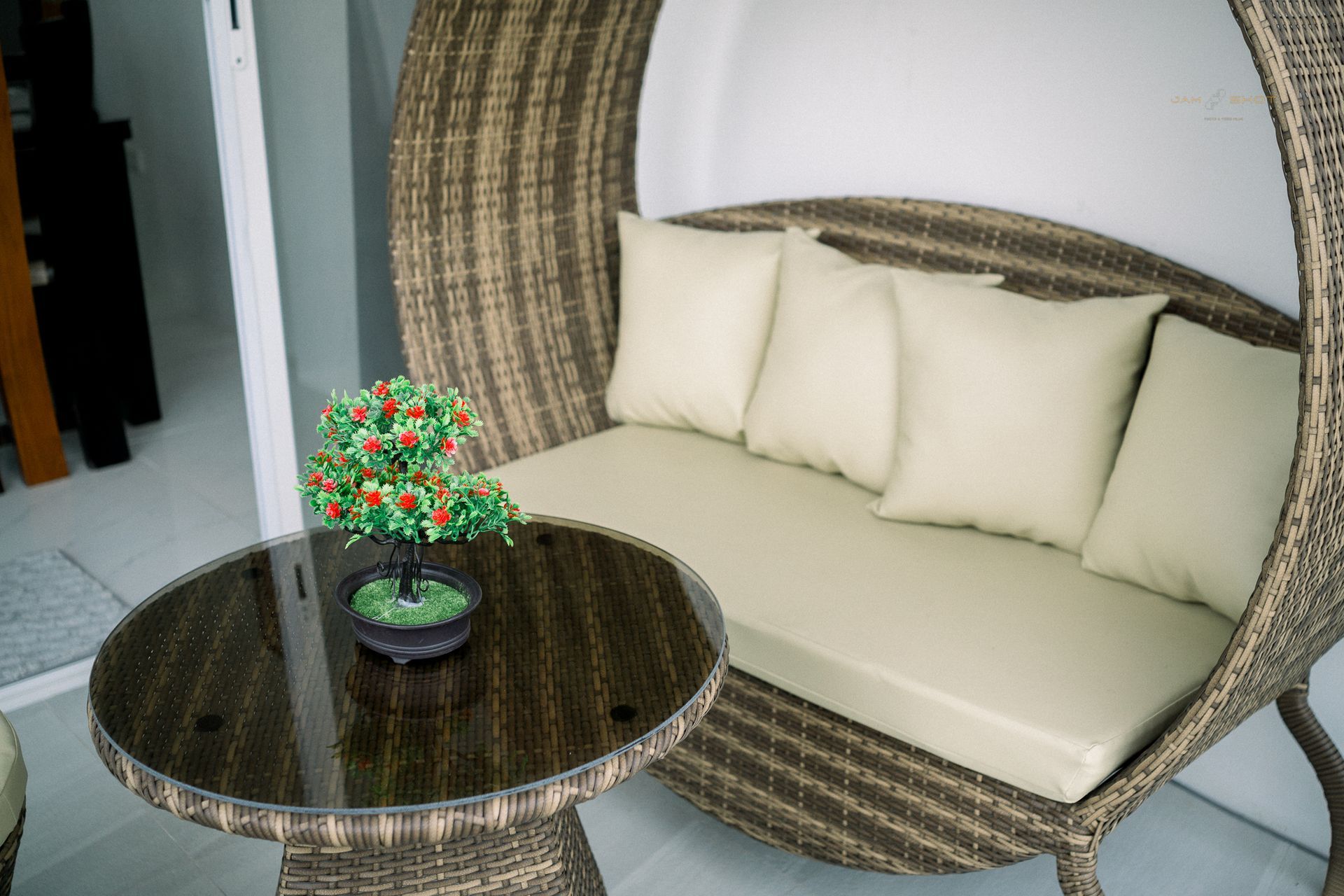 Wicker patio furniture with cream cushions and a glass-top table featuring a small decorative artificial bonsai tree.