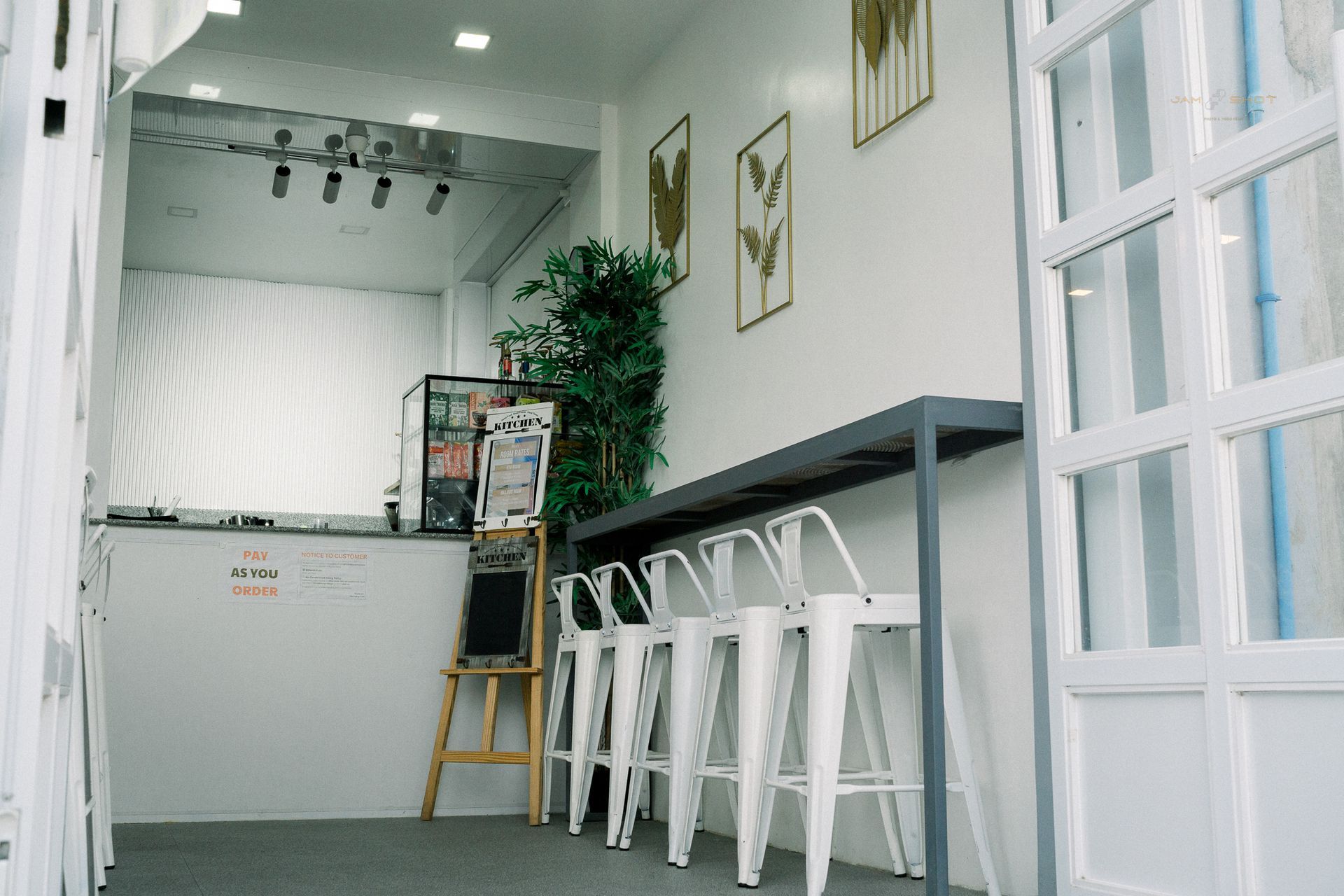A brightly lit, modern minimalist cafe interior with white walls, four white stools at a bar, and framed plant art.