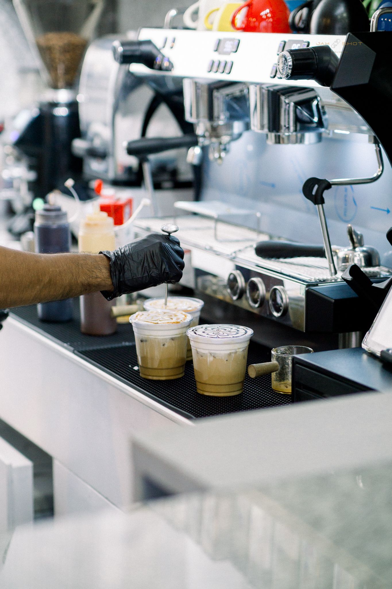 A gloved hand uses a tool to prepare two iced coffee drinks on a counter next to an espresso machine.