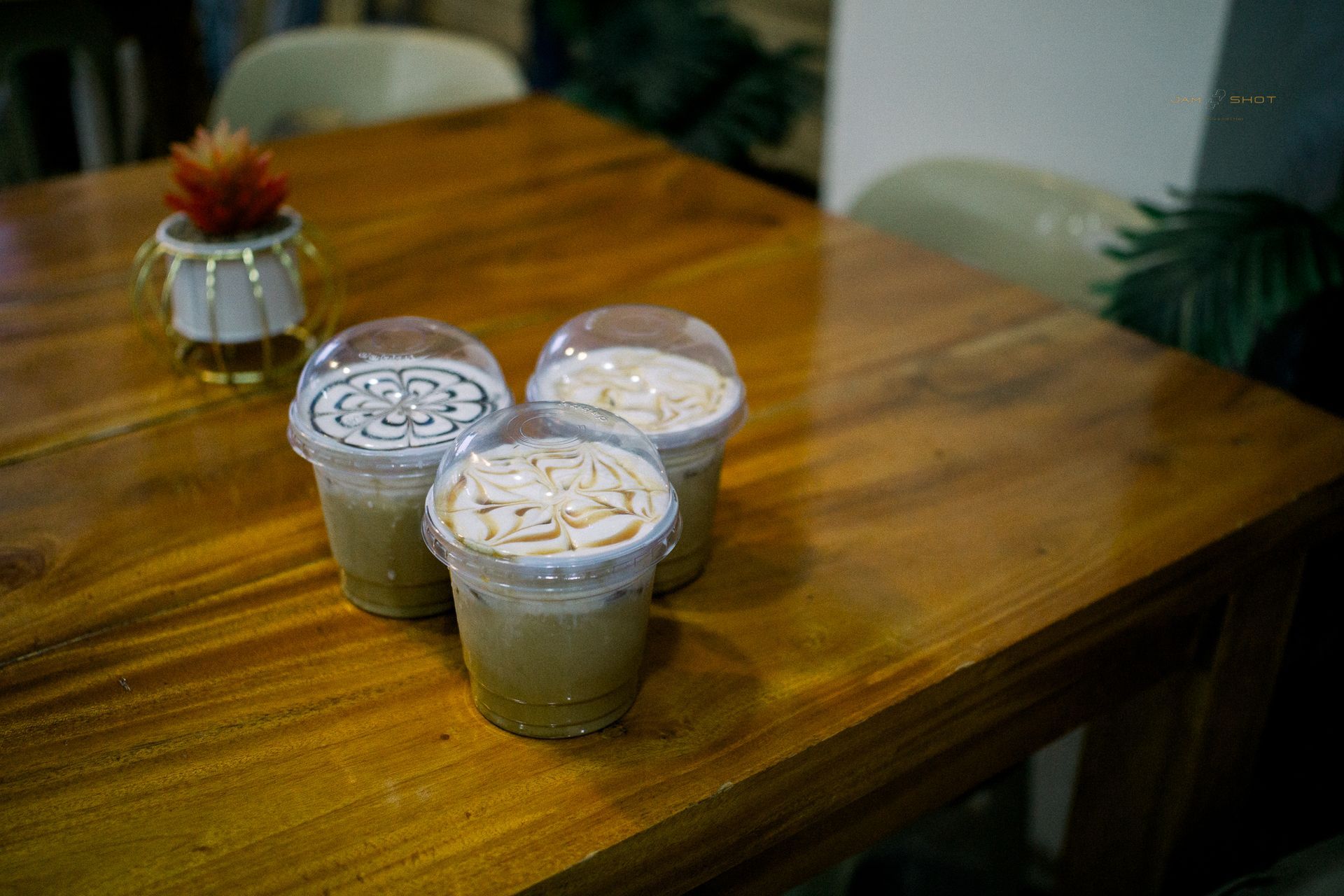 Three clear plastic cups of iced coffee with whipped topping and chocolate drizzle sit on a wooden table.
