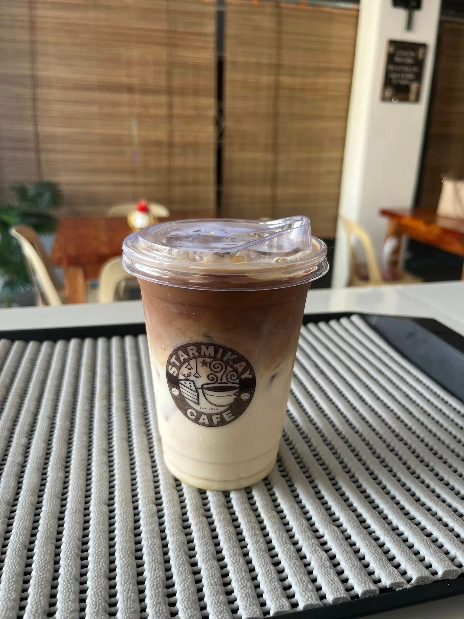 A clear plastic cup of iced layered coffee with a cafe logo, resting on a textured gray mat in a cafe setting.