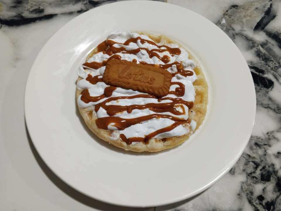 A waffle topped with whipped cream, caramel drizzle, and a Lotus Biscoff cookie on a white plate over a marble table.