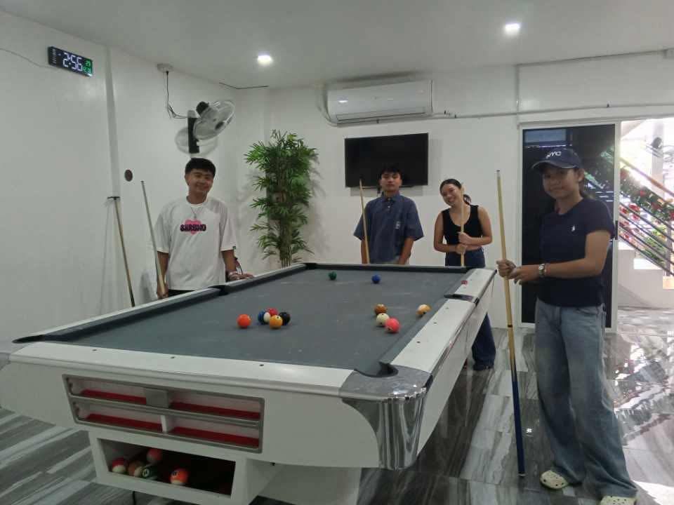 Four people pose around a gray pool table in a bright, modern room, some holding cues and smiling.