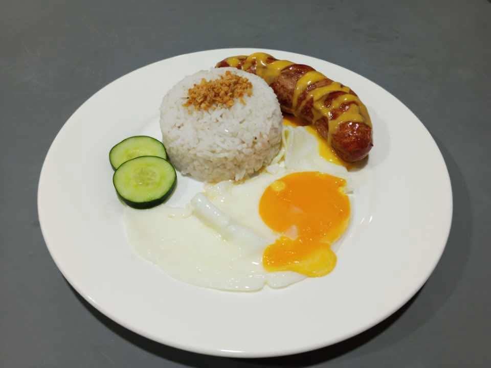 A plate with a scoop of rice topped with crispy garlic, a fried sausage with mustard, a sunny-side up egg, and cucumber.