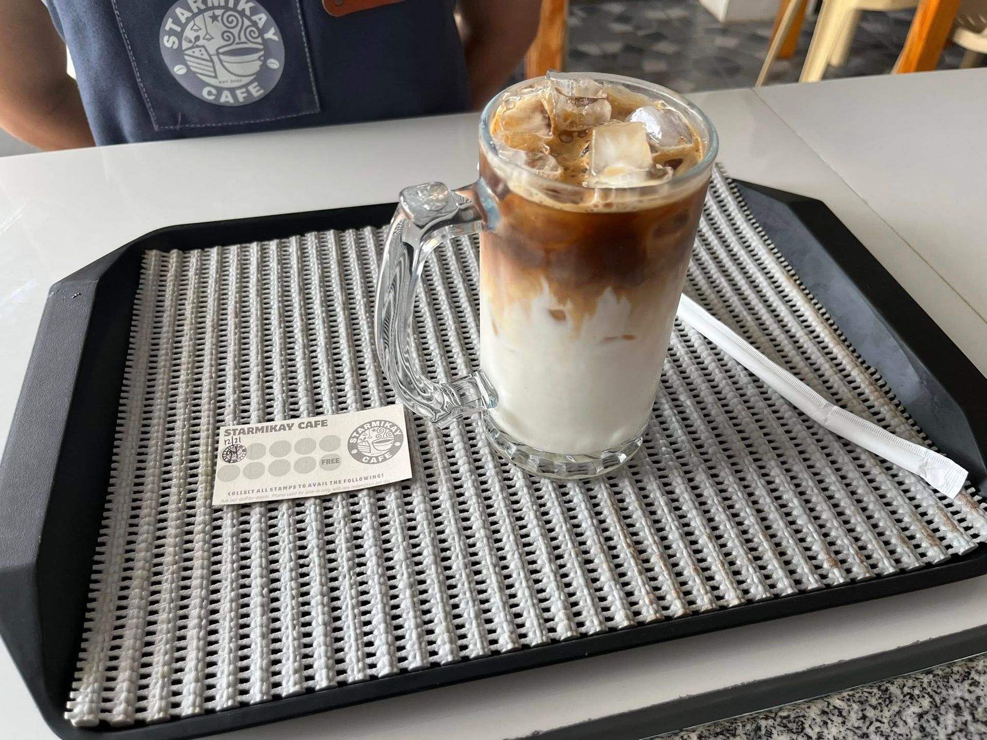 A glass mug of iced coffee with milk on a black tray with a straw and a loyalty card, set on a cafe table.